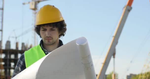 Concentrado jovem construtor em capacete amarelo sobre fundo de construção inacabado. Arquiteto caucasiano analisando desenho. Ao ar livre . — Vídeo de Stock