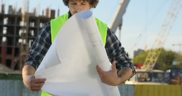 Joven constructor guapo en casco amarillo sobre fondo de construcción sin terminar. Arquitecto analizando dibujo y mirando al costado. Exterior . — Vídeo de stock