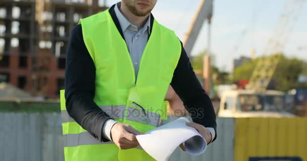 Joven constructor caucásico en casco blanco sobre fondo de construcción inacabado sosteniendo el dibujo y mirando a la cámara. Exterior . — Vídeo de stock