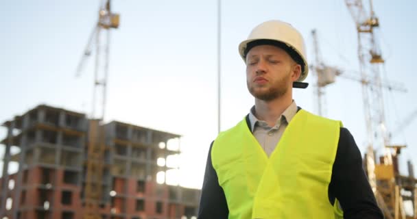 Beau architecte caucasien en casque blanc debout sur le fond du bâtiment. Constructeur caucasien regardant dessin et sourire. Extérieur . — Video