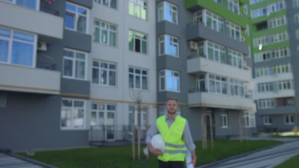 Unga builder med svart lockigt hår håller i hand vita hjälmen och kommer att kameran på bakgrunden byggt hus. — Stockvideo