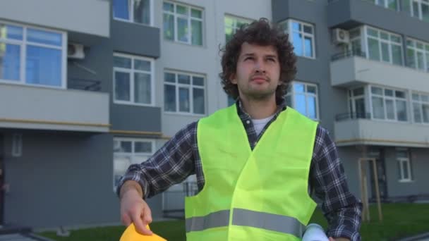 Joven constructor caucásico con el pelo rizado negro sosteniendo en la mano el dibujo, poniendo en la cabeza el casco, componiendo las manos. Exterior . — Vídeos de Stock