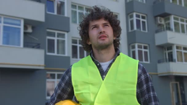 Bonito jovem construtor com cabelo encaracolado preto colocando na cabeça o capacete protetor, compondo suas mãos. Ao ar livre . — Vídeo de Stock