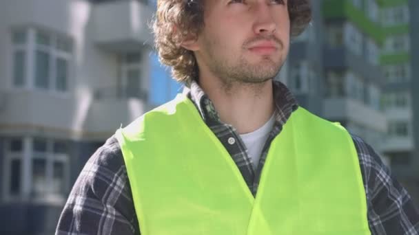 Портрет молодого строителя с черными вьющимися волосами в желтом защитном шлеме, смотрящего на фоне построенного дома . — стоковое видео