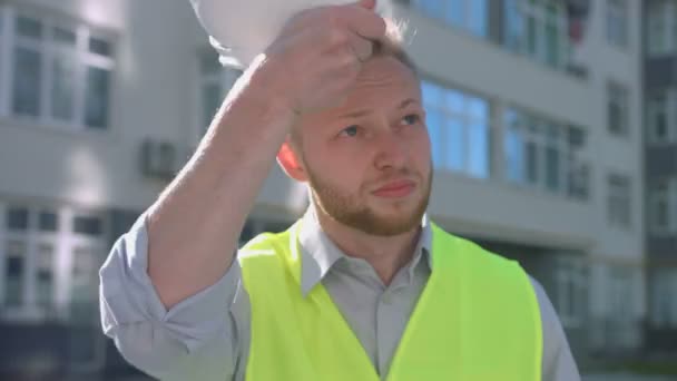 Porträt eines gutaussehenden Bauunternehmers mit rauen Haaren und Bart, der auf dem Kopf einen weißen Schutzhelm aufsetzt. — Stockvideo