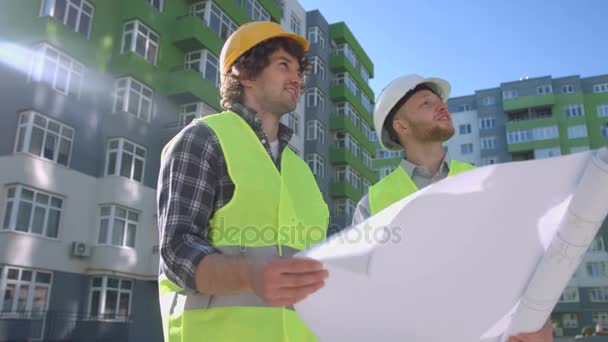 Two architects in protective helmets and green safety jackets, discussing, looking on construction process. Outdoor. — Stock Video