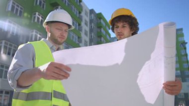 Koruyucu baretler ve yeşil yelek inşaat uygunluğu hakkında konuşurken iki beyaz inşaatçılar çizimi işlemek. Açık.