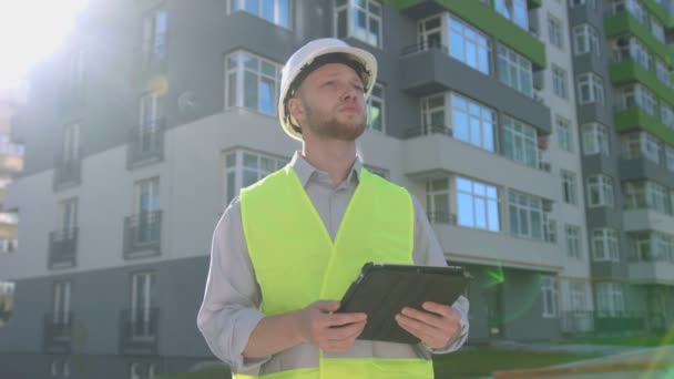 Konzentrierter kaukasischer Bauunternehmer mit weißem Schutzhelm und grüner Sicherheitsweste, der im Hintergrund ein mit Tablets errichtetes Haus für seine Arbeit aufbaut. Außenbereich. — Stockvideo