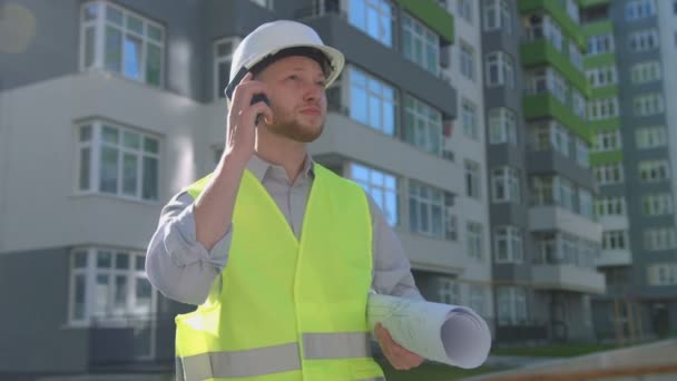 Красивий білий працівник в білому шоломі і зелений жилет розмовляє на мобільному телефоні, стоячи біля побудованого будинку . — стокове відео
