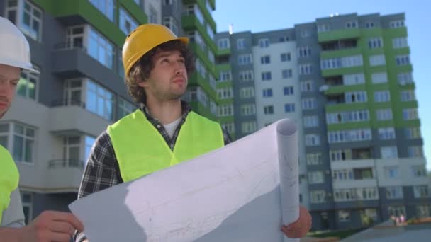 Zwei junge kaukasische Bauarbeiter in Schutzhelmen und Westen, die zeichnend wirken. Arbeiter, der mit dem Handy in der Nähe seines Kollegen steht. Außenbereich. — Stockvideo