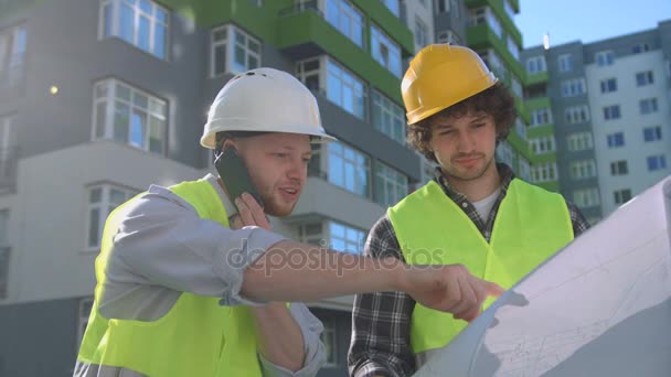 Tým mladých kavkazské stavitelů v chráněných přilby a vesty pohledu výkresu. Stavitel stojí u jeho kolega pomocí chytrého telefonu. Venkovní. — Stock video