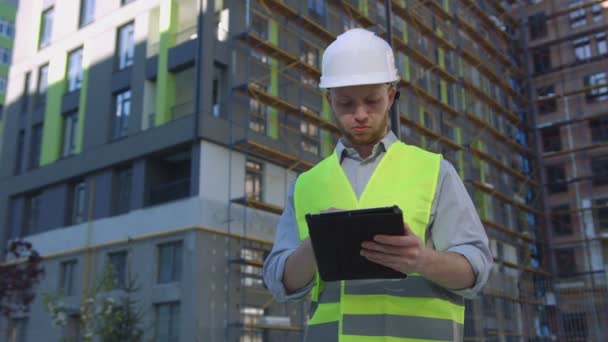 Constructor caucásico serio con casco blanco protegido y chaleco de seguridad verde usando tableta sobre fondo de construcción sin terminar . — Vídeos de Stock