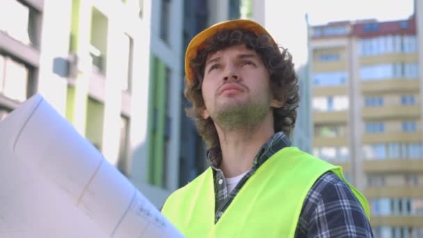 Молодой кавказский строитель в желтом защитном шлеме и жилете оглядывается и держит в руках рисунок. На открытом воздухе . — стоковое видео