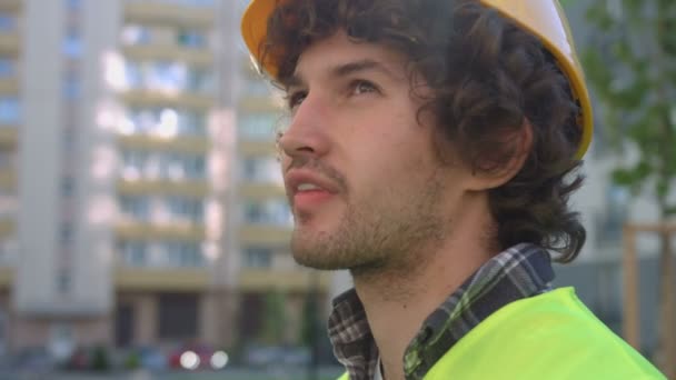 Портрет бокового вида на кавказского молодого строителя с черными вьющимися волосами в защитном шлеме, смотрящего вверх. На открытом воздухе . — стоковое видео