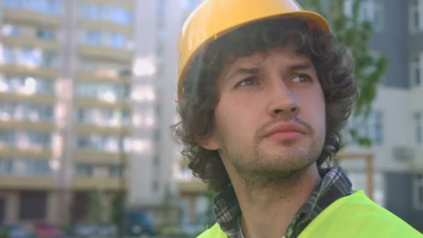 Retrato de joven constructor atractivo con pelo rizado negro en el casco protector, mirando a su alrededor. Exterior . — Vídeos de Stock