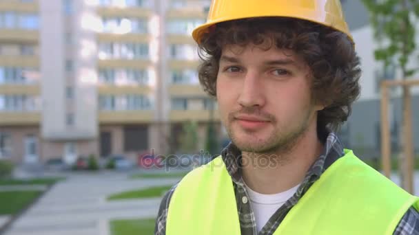 Retrato de joven constructor guapo con el pelo rizado negro en el casco protector posando en la cámara y sonriendo. Exterior . — Vídeos de Stock