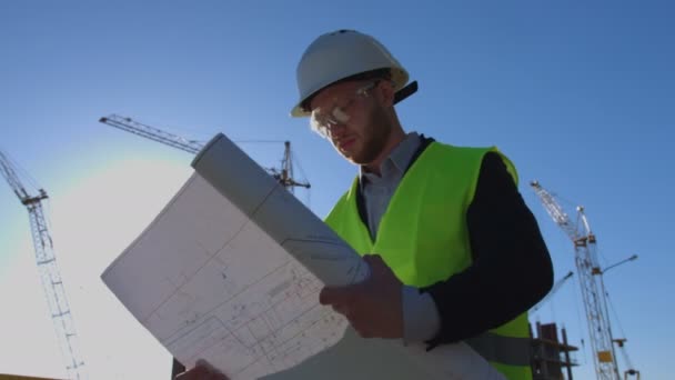 Arquiteto concentrado em óculos protegidos, capacete branco e colete olhando desenho em fundo de construção inacabado . — Vídeo de Stock