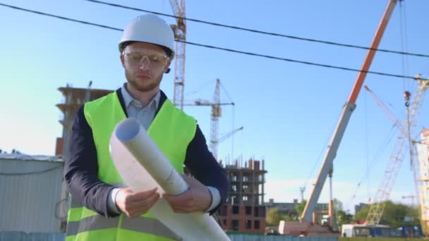 Jeune architecte caucasien en lunettes protégées, casque et gilet regardant dessin et penser à quelque chose sur fond de construction inachevé . — Video