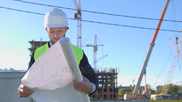 Arquitecto caucásico en gafas protegidas, casco y chaleco desplegando dibujo y analizándolo sobre fondo de construcción inacabado . — Vídeos de Stock