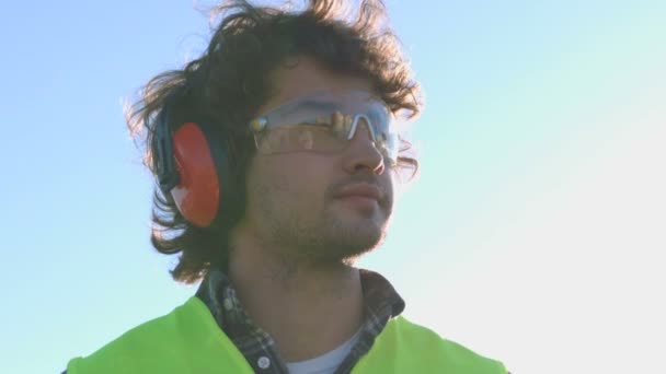 Портрет молодого строителя с черными вьющимися волосами в защитных очках и наушниках. На открытом воздухе . — стоковое видео