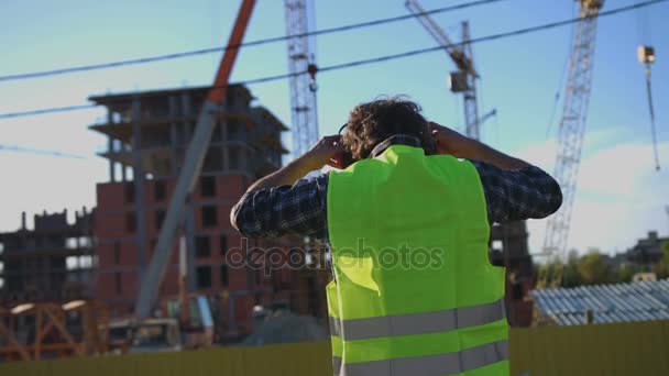 Vue arrière du constructeur avec des cheveux bouclés noirs en gilet vert mettant sur la tête les écouteurs sur fond de construction inachevé . — Video