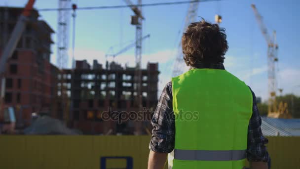 Вид ззаду будівельника з чорним кучерявим волоссям у зеленому жилеті, що стоїть на незакінченому будівельному тлі . — стокове відео