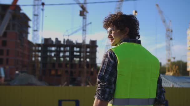 Vista posterior del constructor con el pelo rizado negro en chaleco verde mirando en el lado, poniendo en la cabeza el casco sobre fondo de construcción sin terminar . — Vídeos de Stock