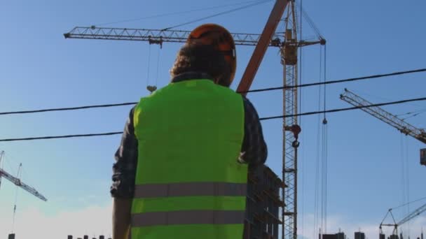 Zadní pohled na tvůrce s černými kudrnatými vlasy v košili a zelené vesty při pohledu na proces výstavby, nasadil do hlavy žlutou helmu. Venkovní. — Stock video