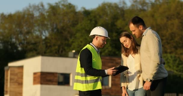 Ein kaukasischer Architekt mit weißem Helm zeigt jungen Bauherren auf einem Tablet die Zeichnung eines Hauses. Außenbereich. — Stockvideo