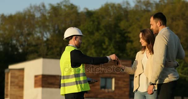 Architecte caucasien en casque blanc serrant la main du client et de la femme souriant et embrassant son mari. Extérieur . — Video