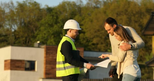 Кавказский архитектор в белом шлеме с мужчинами и женщинами-клиентами обсуждает строительный проект с использованием чертежа и планшета. На открытом воздухе . — стоковое видео