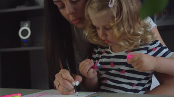 Очаровательная маленькая девочка со светлыми вьющимися волосами и ее мать с длинными черными волосами рисует с войлочной ручкой на листе бумаги. В помещении . — стоковое видео