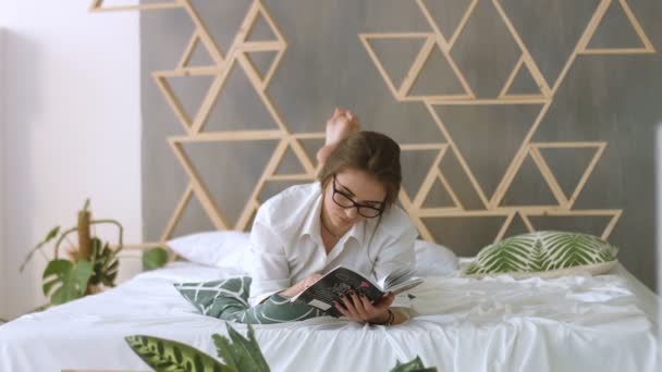 Junge schöne Frau mit Brille bereitet sich auf Prüfungen vor, während sie auf dem Bett gelyncht wird. — Stockvideo