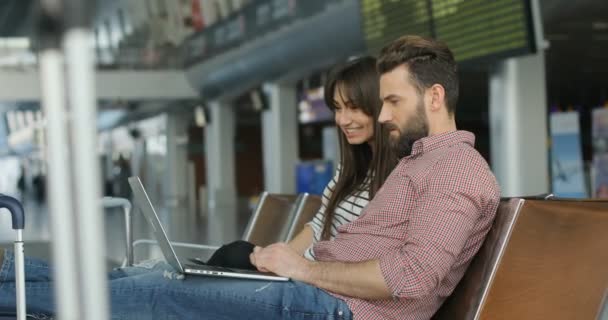 Retrato de feliz joven pareja amorosa beata está sentado en el salón del aeropuerto, el hombre usando el ordenador portátil y mostrando algo a la mujer. Vista lateral . — Vídeo de stock