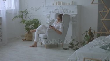 Genç çekici kadın sandalyede oturma odasında otururken tablet kullanarak beyaz casual giyim.