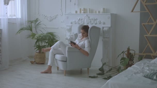 Jeune femme attrayante en blanc vêtements décontractés en utilisant la tablette. assis dans la chaise dans le salon . — Video