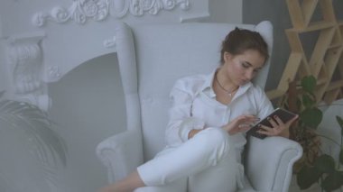 Güzel beyaz genç kadın evde oturma odasında sandalyede oturan ve tablet kullanma.