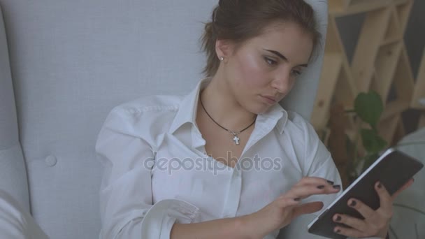 Porträt einer jungen attraktiven Frau, die zu Hause im Wohnzimmer auf einem Stuhl sitzt und ein Tablet benutzt. — Stockvideo