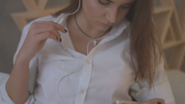 Jovem mulher de camisa casual ouvindo música em fones de ouvido enquanto deitada na cama em casa . — Vídeo de Stock