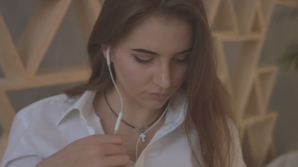 Mujer joven en camisa casual escuchando música en auriculares mientras se relaja en la cama en casa . — Vídeos de Stock