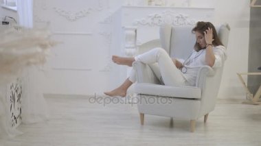Evde sandalyede otururken akıllı telefon kullanan genç kadın rahat gömlek dinleme müzik kulaklık,.