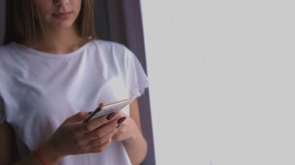 Ung kvinna med hjälp av den smarta telefonen för chatta med vänner, stående nära fönstret hemma. — Stockvideo