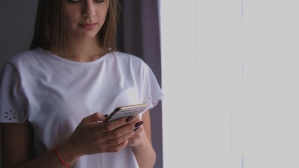 Молода жінка дивиться у вікно і використовує смартфон для спілкування . — стокове відео