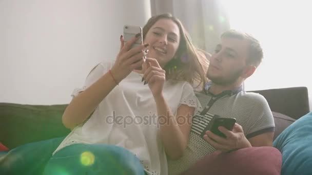 Mujer joven que viene a la sala de estar, sentado en el sofá y mostrando algo divertido en el teléfono inteligente a su novio . — Vídeos de Stock