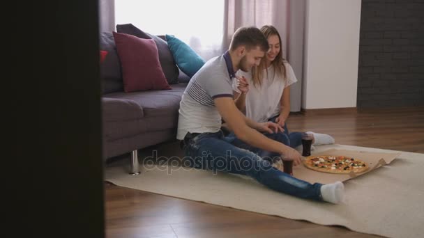 Joven feliz hombre y mujer sentados en el suelo en casa y comiendo sabrosa pizza. Vista desde el costado . — Vídeos de Stock