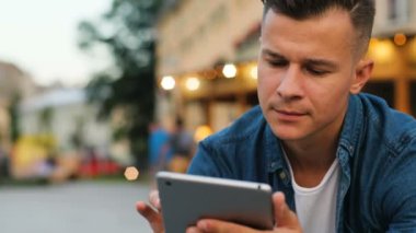Şehir merkezindeki bankta otururken tablet kullanarak rahat tişörtlü yakışıklı adam portresi. Çekim açık.