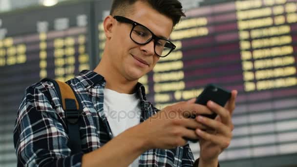 Close-up shot van jonge man met bril met behulp van slimme telefoon voor het controleren van zijn vlucht op de achtergrond van de tabel aankomsten glimlachen. — Stockvideo