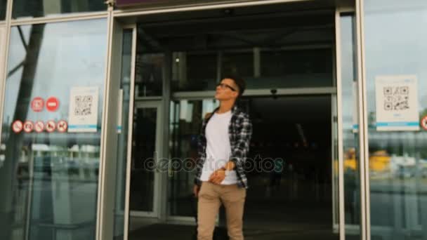 Young handsome man in the glasses with the baggage walking from the door of modern airport terminal to his home. Dolly shot. — Stock Video