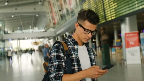 Porträt eines jungen Mannes mit Brille und Gepäck, der im modernen Flughafenterminal spazieren geht und das Smartphone zum Chatten benutzt. — Stockvideo