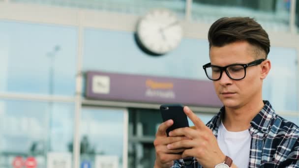 スマート フォンを使用して近代的な空港の背景で友達と話しているガラスのスタイリッシュな若者。クローズ アップ. — ストック動画